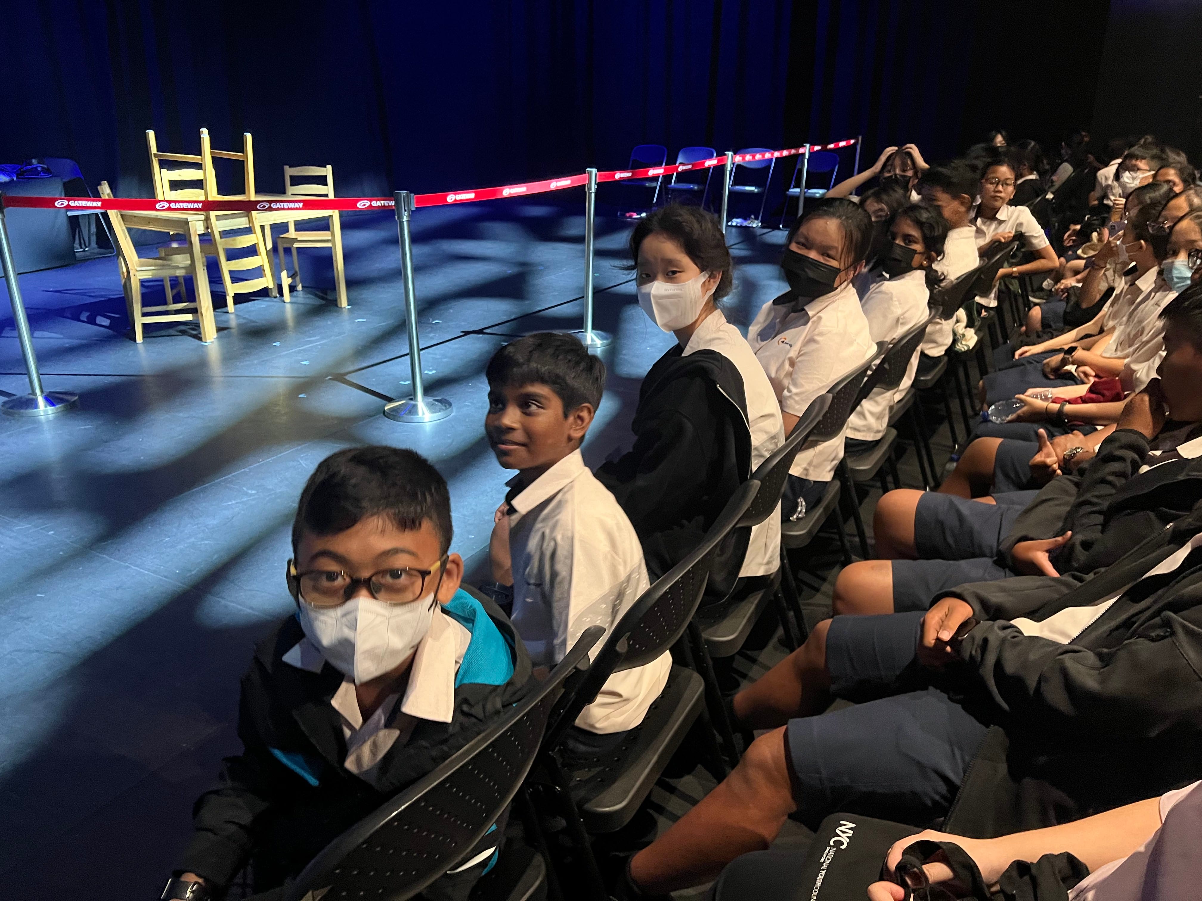 School children in uniforms and face masks sit in rows of chairs beside a blue-lit stage with wooden chairs.